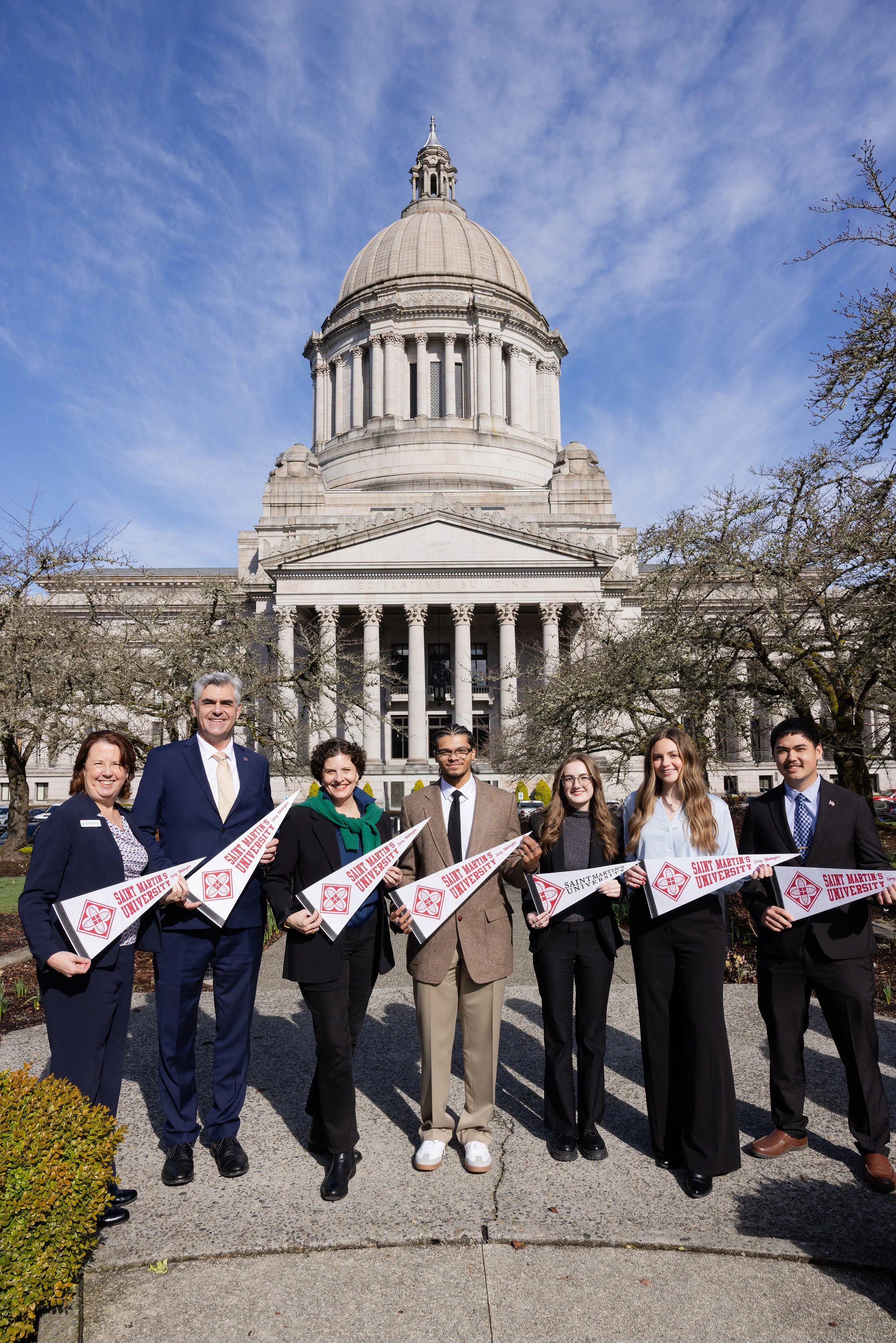 Group of seven diverse young adults and two older adults standing in front of a large government building with a dome, holding flags that read "Saint Martin's University," outdoors on a clear day with blue sky.