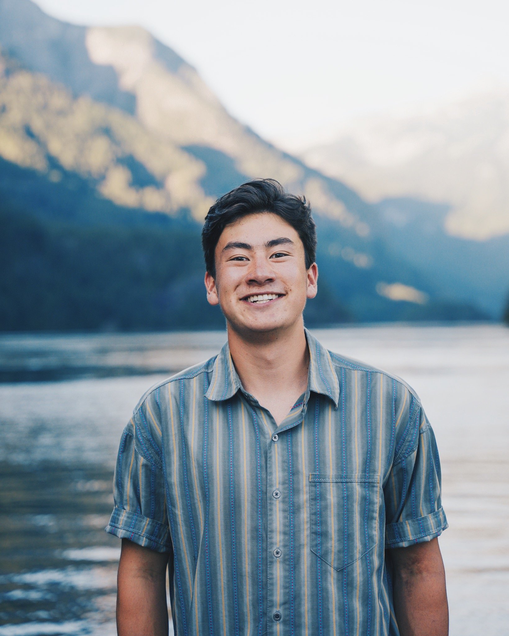 A young man smiling outdoors by a river, with mountains in the background.