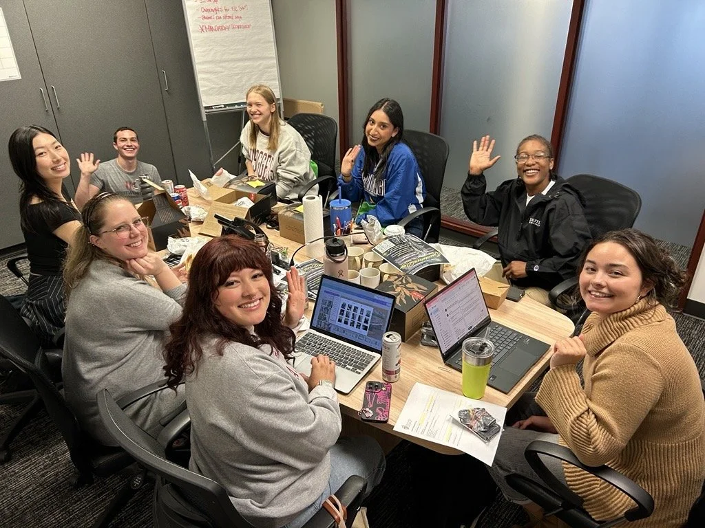 A group of nine women gathered around a meeting table, smiling and waving at the camera in a casual office setting.