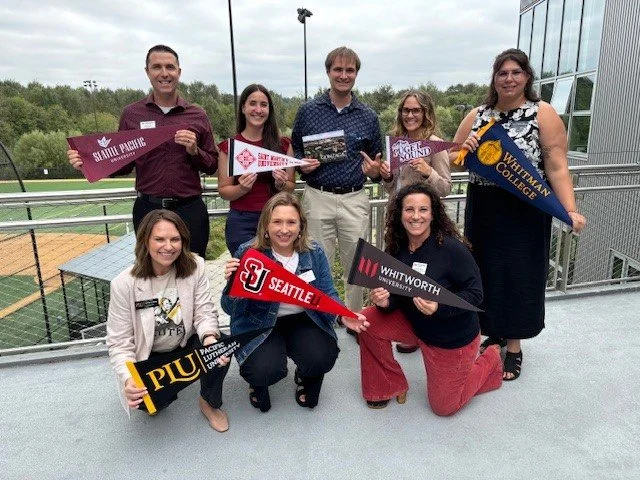 Group of seven people holding college banners and signs, standing and kneeling on a balcony overlooking a sports field with trees in the background.