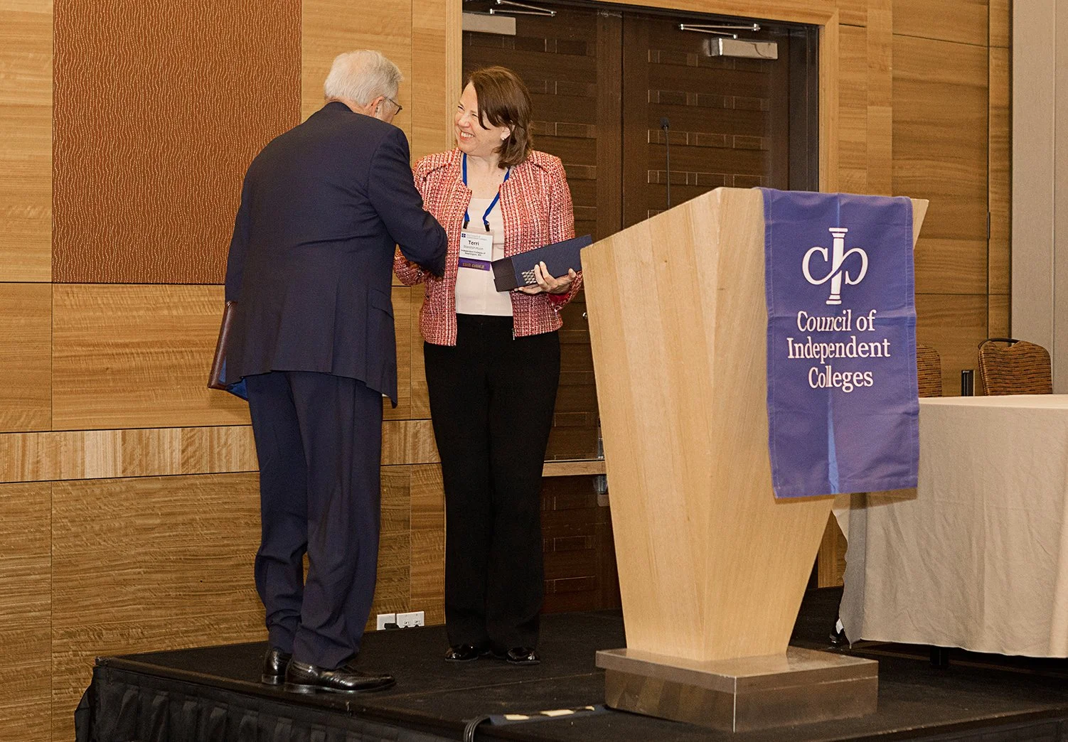 A man and woman shaking hands on a stage with a wooden podium that has a Council of Independent Colleges banner, in a conference room setting.