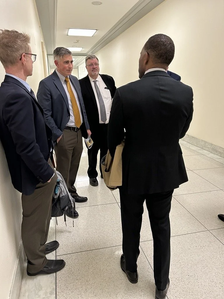 Four men in business suits having a conversation in a hallway, one man facing away from the camera, while the other three are facing him.