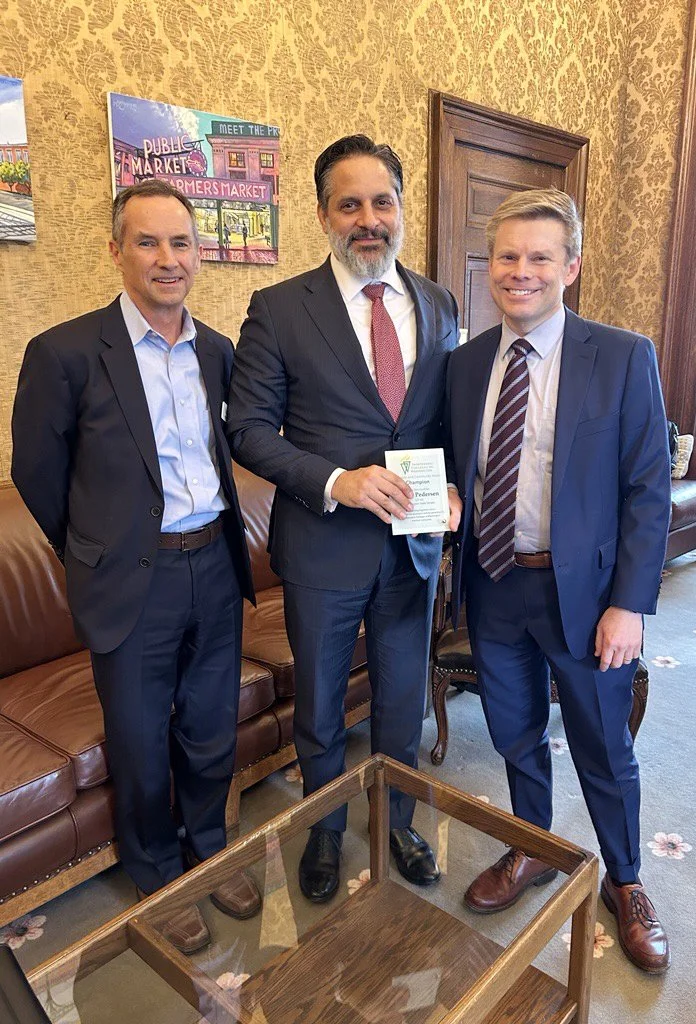 Three men in business suits standing together in a room with patterned wallpaper. The man in the middle is holding a certificate. They are smiling and standing near a leather couch and a wooden table.