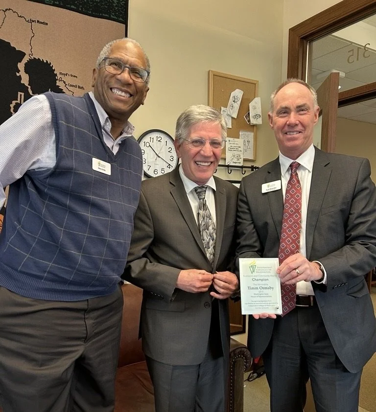 Three men in suits and a man in a vest smiling for a photo, with one holding a certificate, inside an office with a wall clock and bulletin board in the background.
