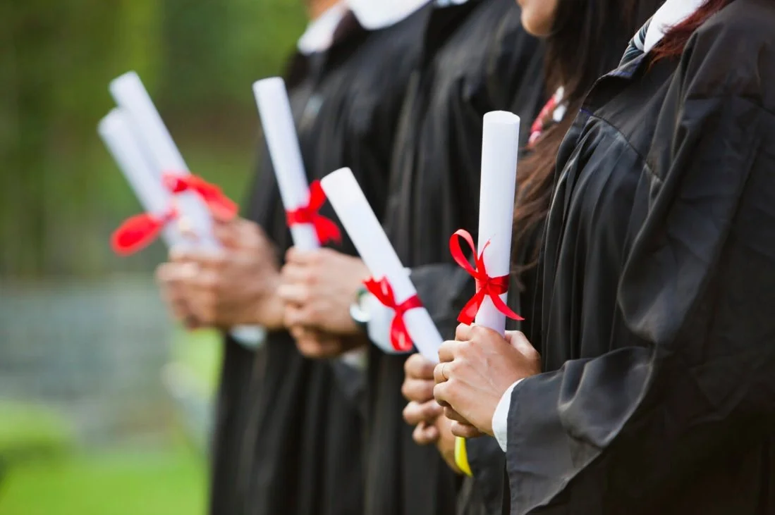 Graduates wearing black gowns holding diplomas wrapped with red ribbons during a graduation ceremony.