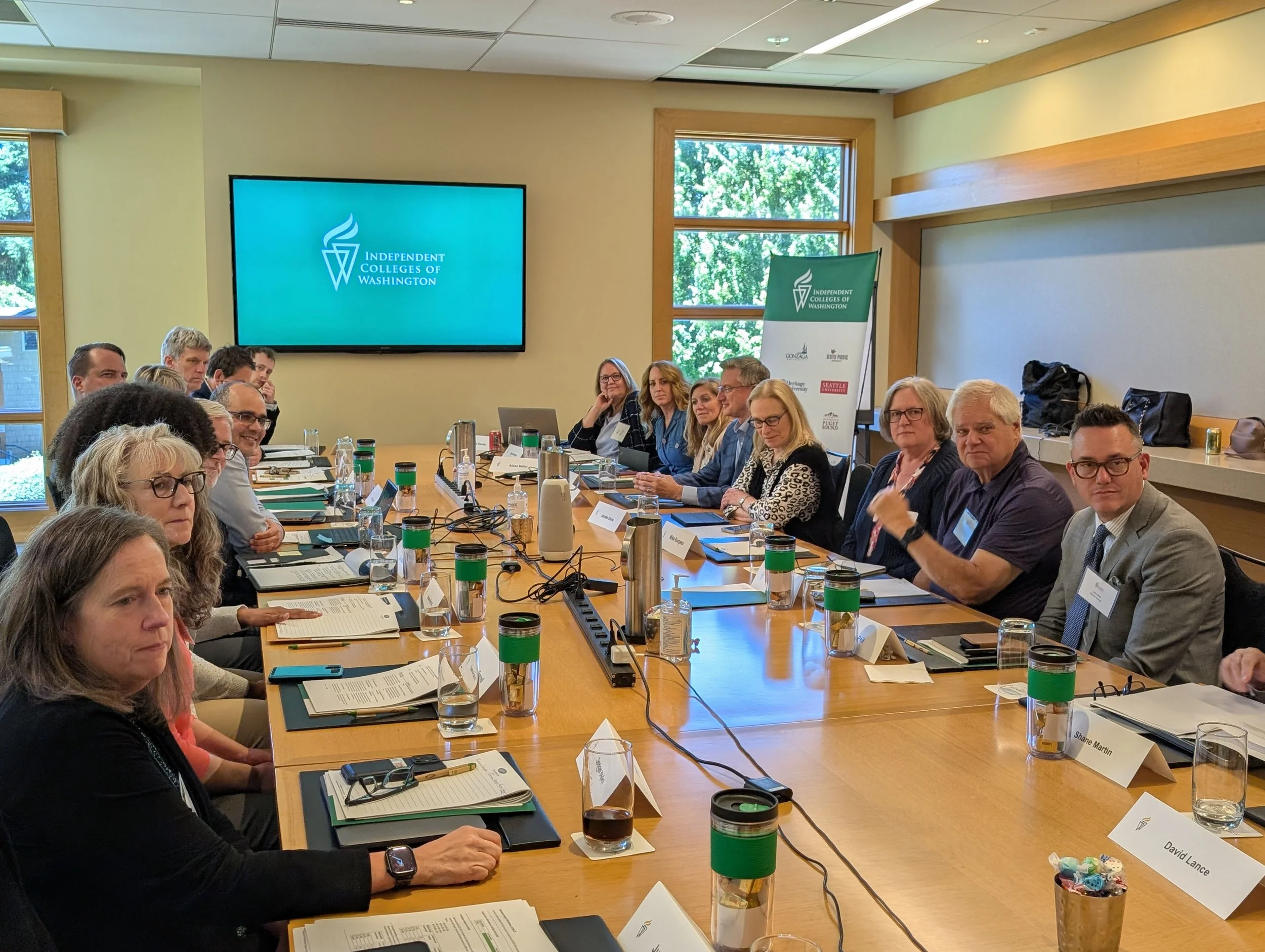 A group of people sitting around a conference table at a meeting, with a large monitor displaying the logo of the Independent Colleges of Washington.