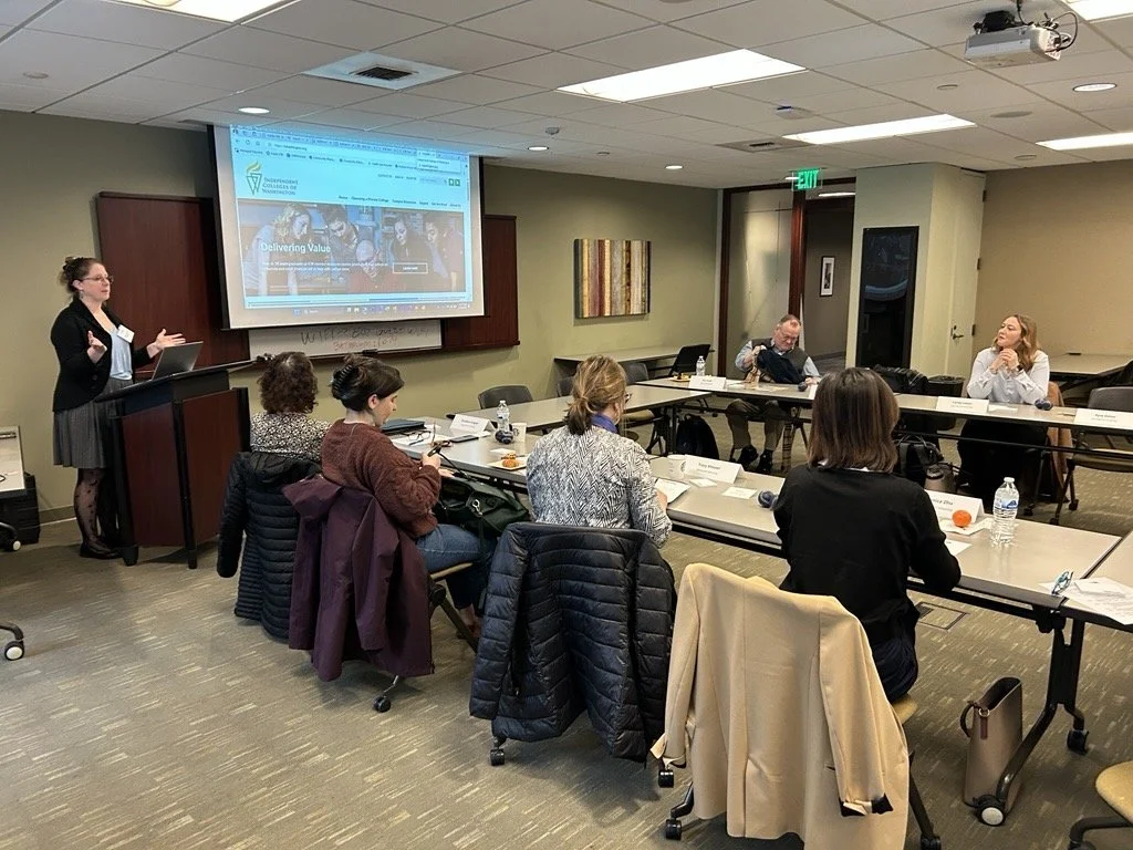 A woman standing at a podium speaking to a group of people sitting around a U-shaped conference table in a meeting room.