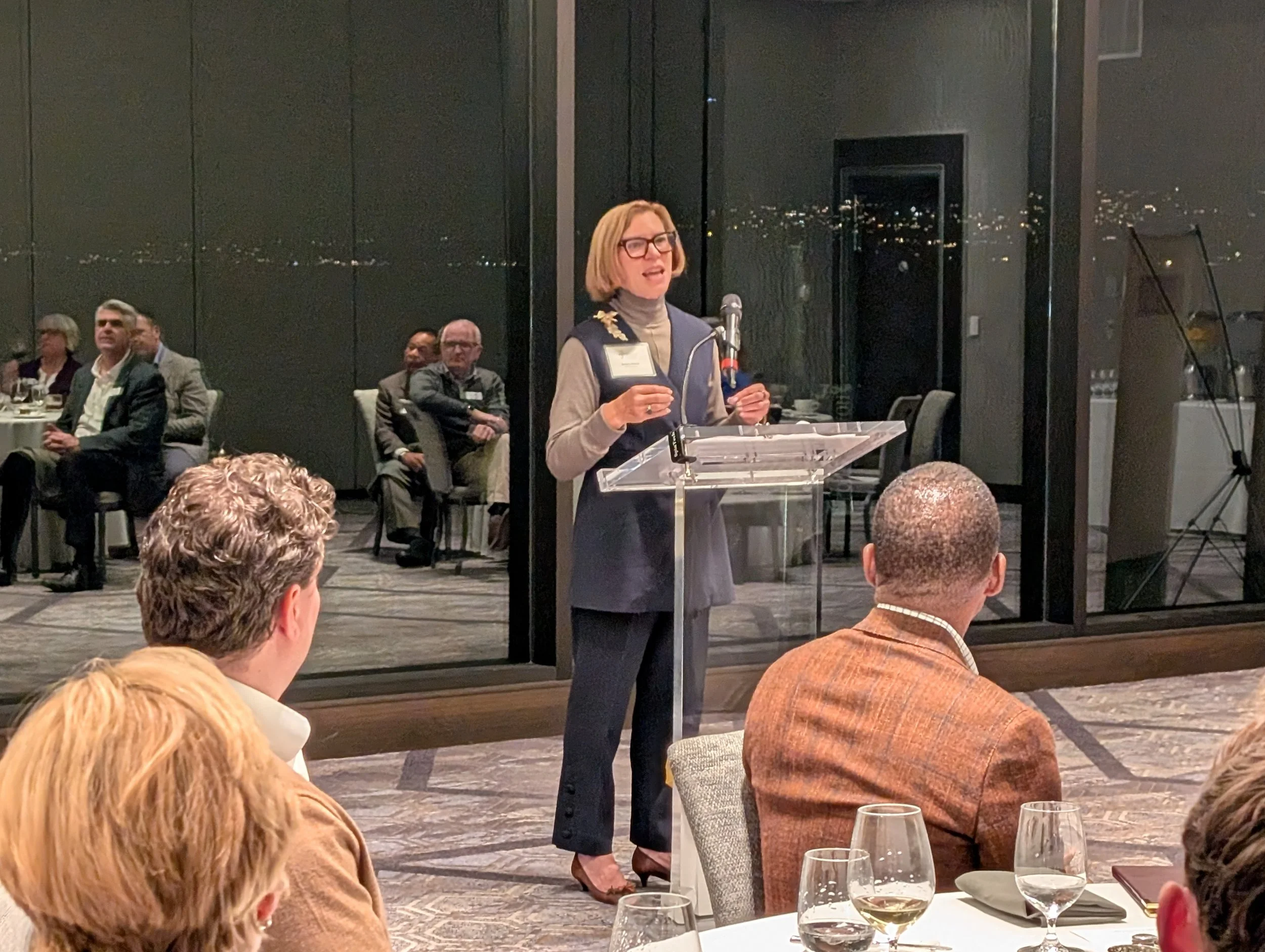 Woman speaking at a podium during a formal event, with seated attendees watching inside a room with glass walls and city view at night.