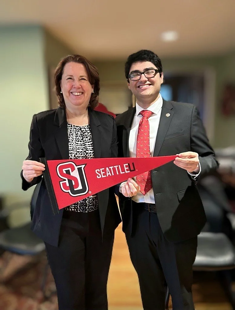 Two smiling people, a woman and a man, holding a red Seattle University pennant. The woman is wearing a black blazer and patterned blouse, and the man is dressed in a grey suit with a red tie. They are standing indoors in a warmly lit room.