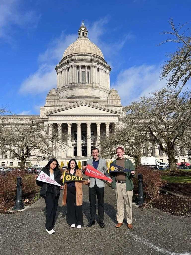 Four students standing in front of a large domed government building, holding signs for colleges and universities, with trees and parked cars in the background.