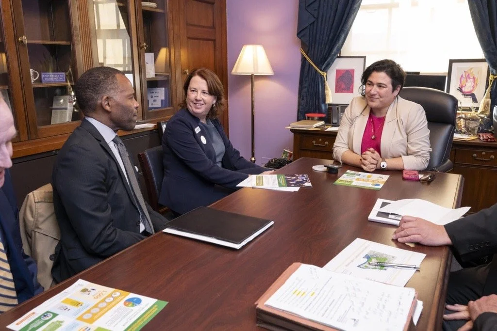 A woman in a beige blazer sitting at a desk and conversing with three people across from her, in an office with wooden furniture and framed artwork on the wall.