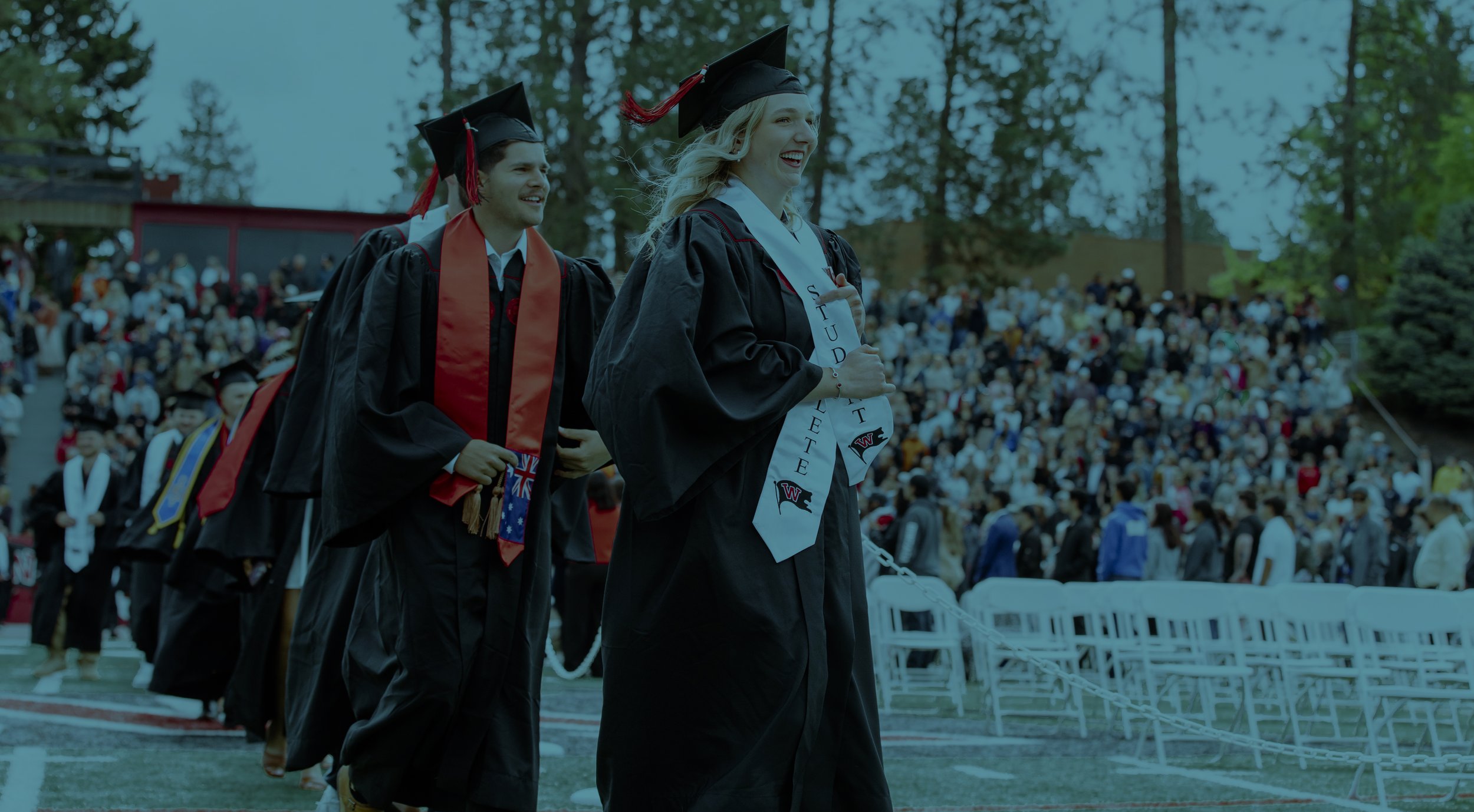 Group of diverse graduates in caps and gowns attending a graduation ceremony, smiling and wearing various cords and leis.