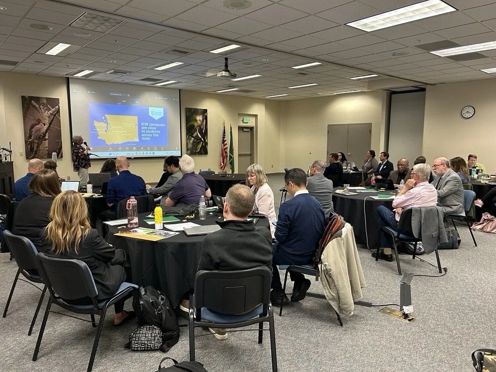 A conference room filled with attendees seated around black tables, listening to a presenter at the front and looking at a large screen displaying a presentation, with flags on the side and a clock on the wall.