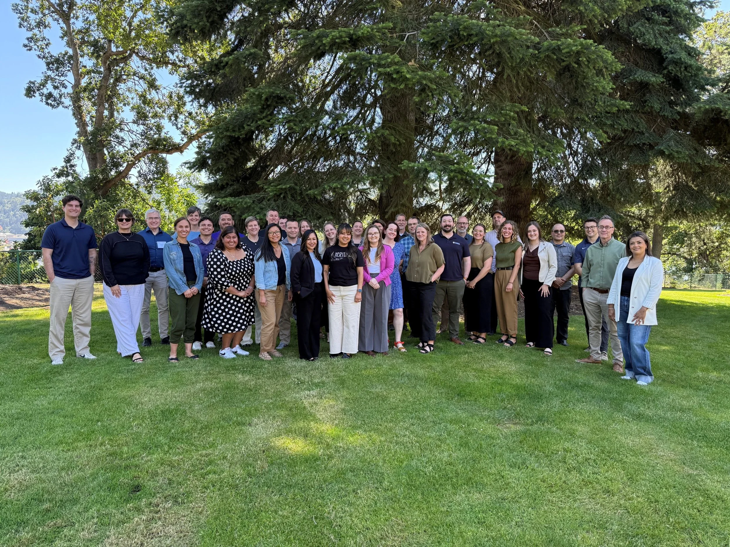 A large group of diverse people standing outdoors on grass in front of a large tree with green leaves, with mountains and blue sky in the background.