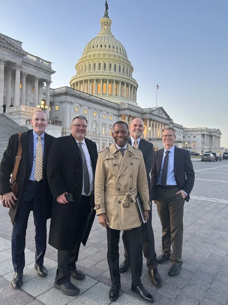 Group of five men standing in front of the U.S. Capitol building, smiling, dressed in business attire and a trench coat, during daytime.