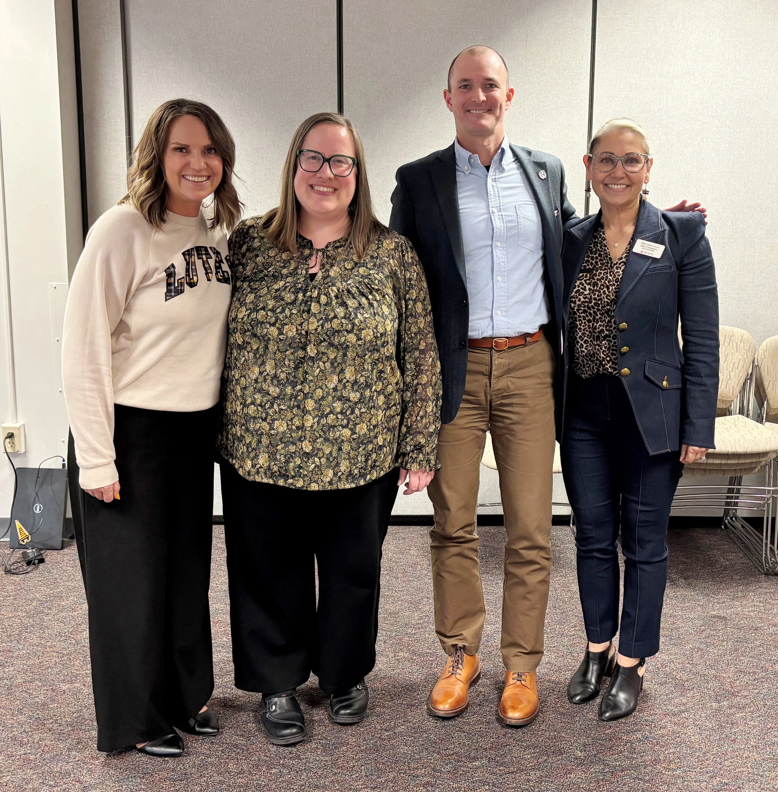 Four women and one man standing together in an indoor setting, smiling and posing for the photo. They are dressed in semi-formal to business casual attire.