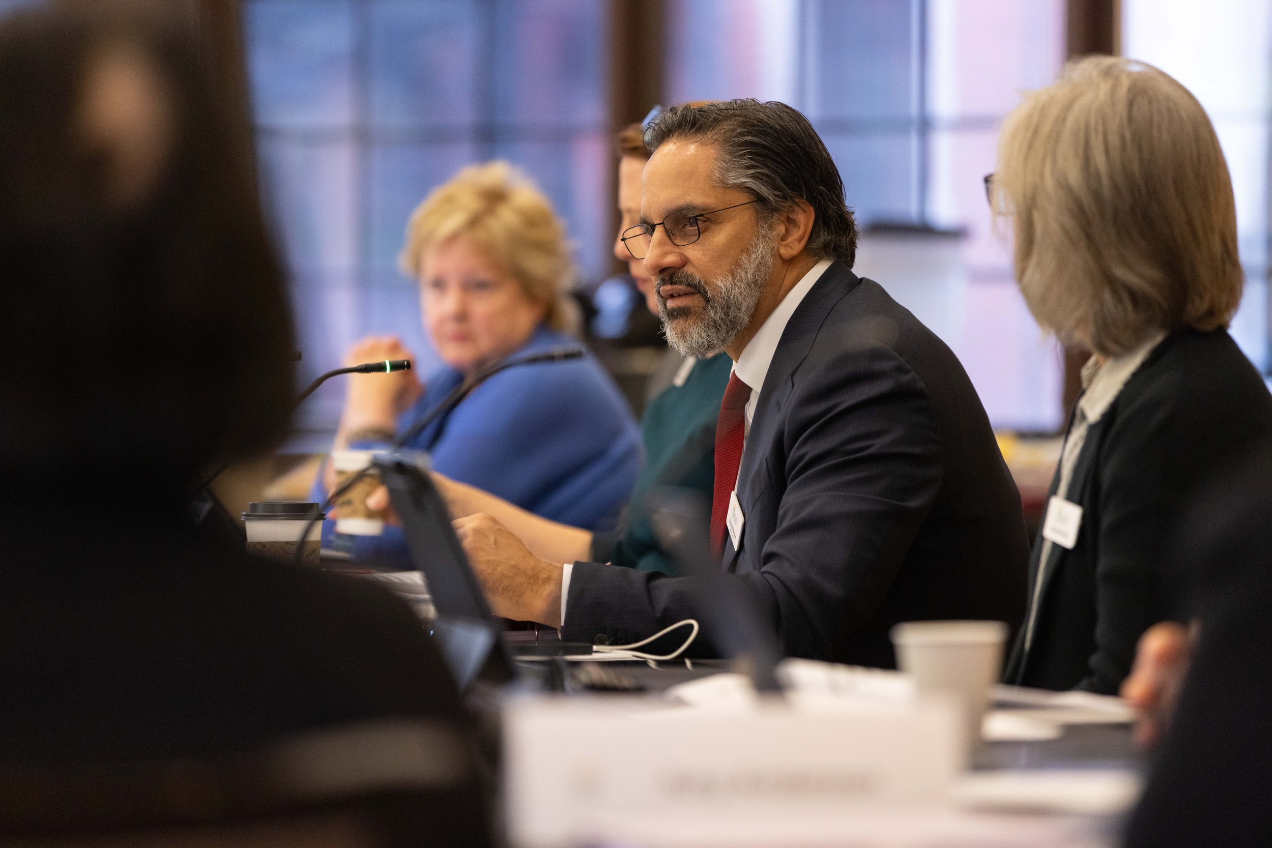 A group of professionally dressed individuals seated at a conference table, engaged in a discussion, with a focus on a man with glasses and a beard.