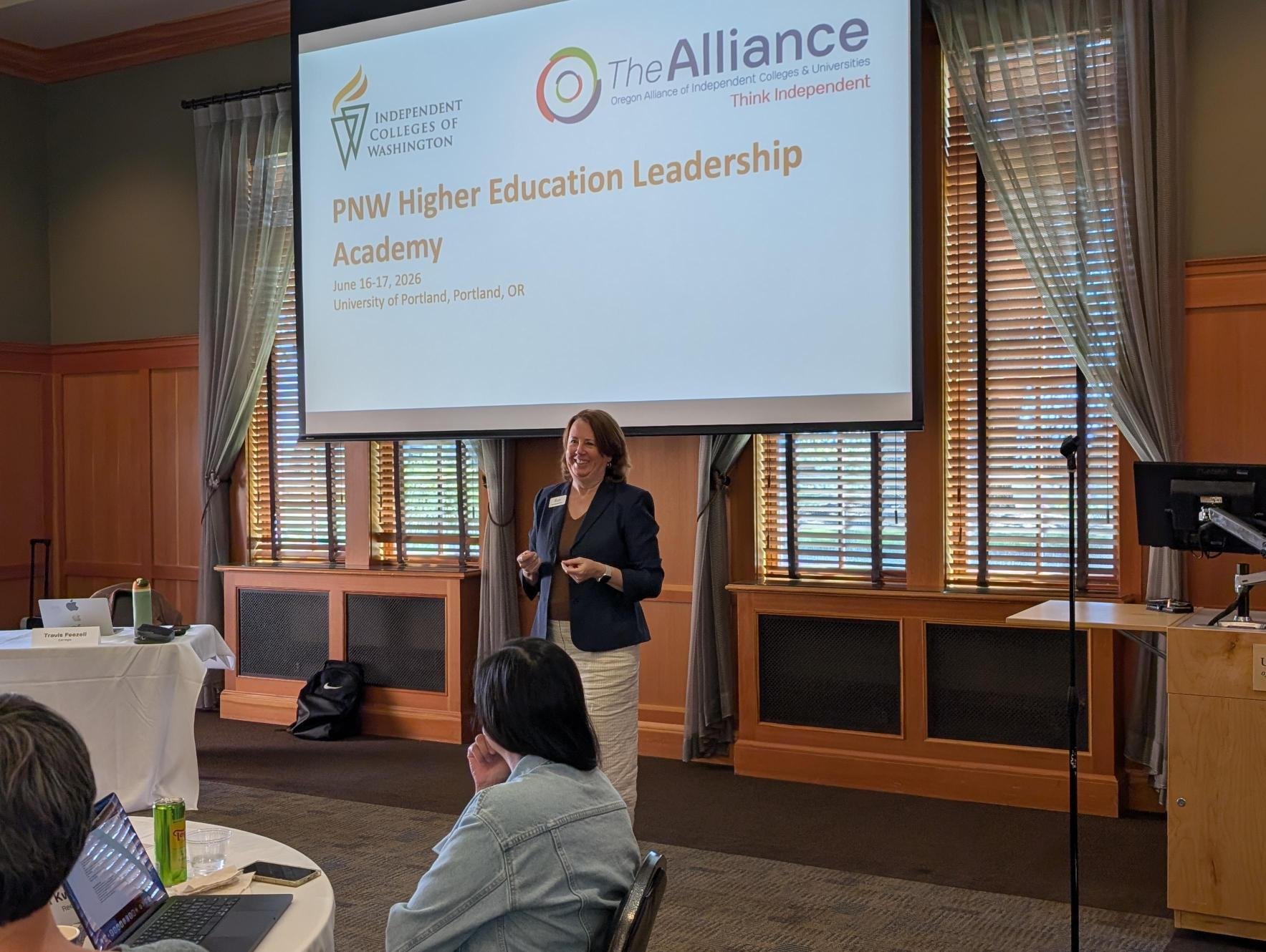A woman speaking at a presentation during the PNW Higher Education Leadership Academy, June 16-17, 2026, at the University of Portland, Portland, Oregon. The presentation slide displays logos for Independent Colleges of Washington, Oregon Alliance of Independent Colleges & Universities, and the event details.