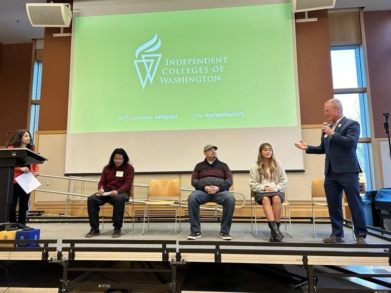 A panel discussion at the Independent Colleges of Washington event with four people on stage, including a man in a suit speaking into a microphone, and a large screen behind them displaying the ICCW logo and Wi-Fi info.