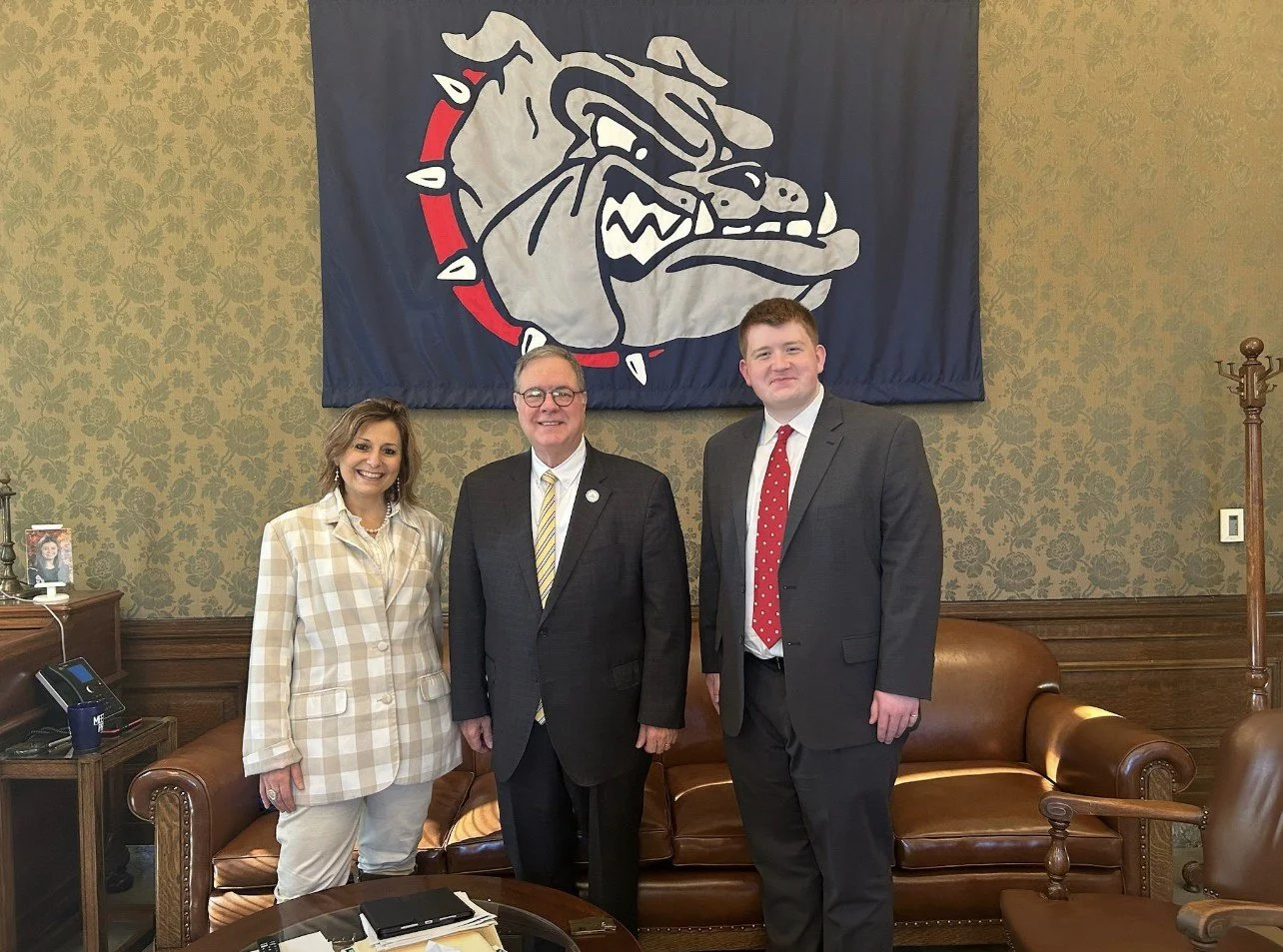 Three people standing in front of a Gonzaga University banner in a room with wood paneling and floral wallpaper. The group includes a woman on the left and two men in suits, all smiling.