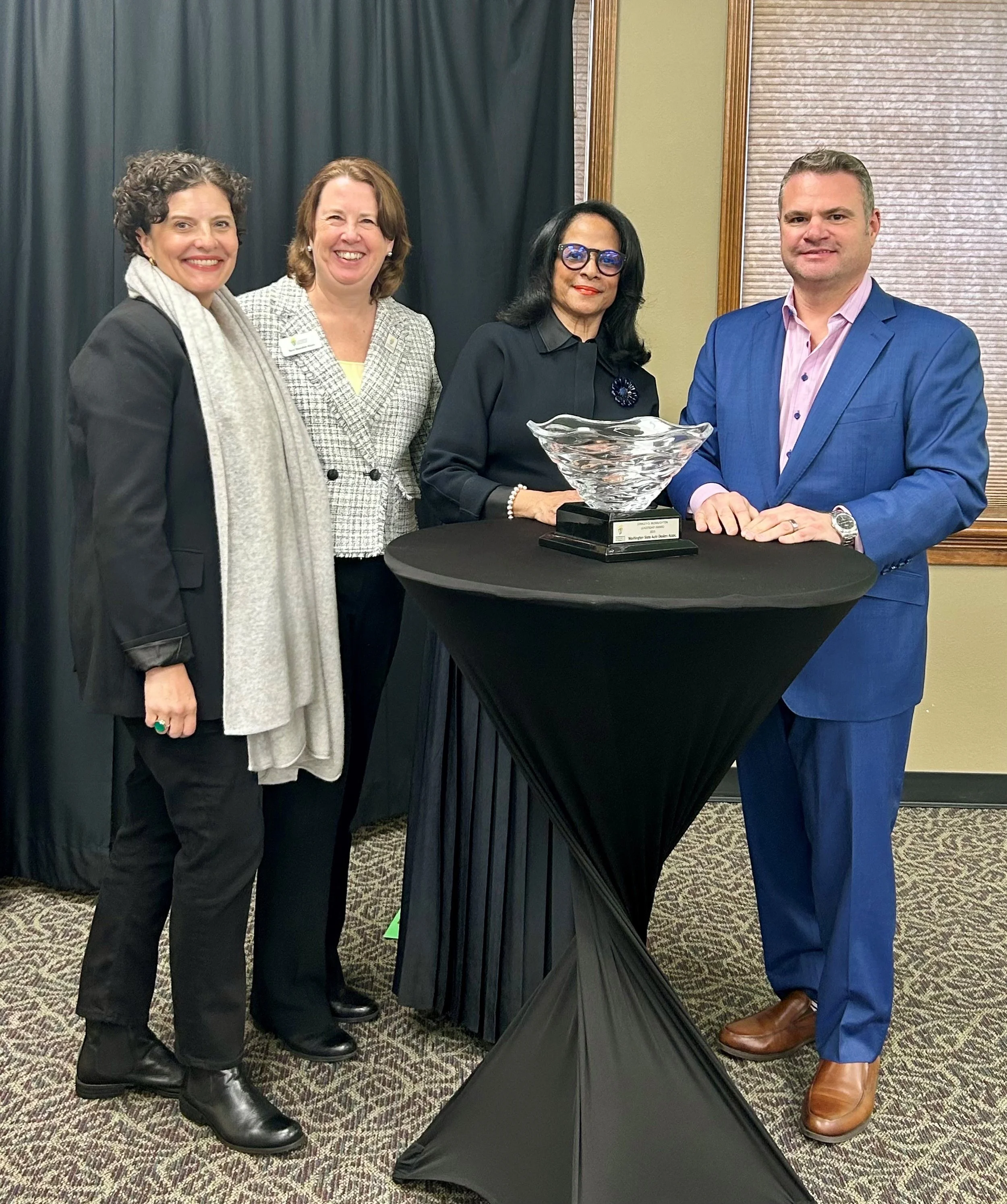 Four people standing behind a black table with a clear glass bowl on it. They are dressed in business attire, posing for a photo in an indoor setting with a dark curtain and a window with blinds in the background.
