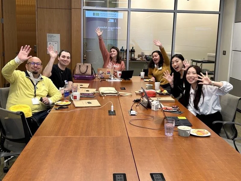 Six people sitting around a conference table, smiling and raising their hands in a meeting or celebration, with snacks and beverages on the table.