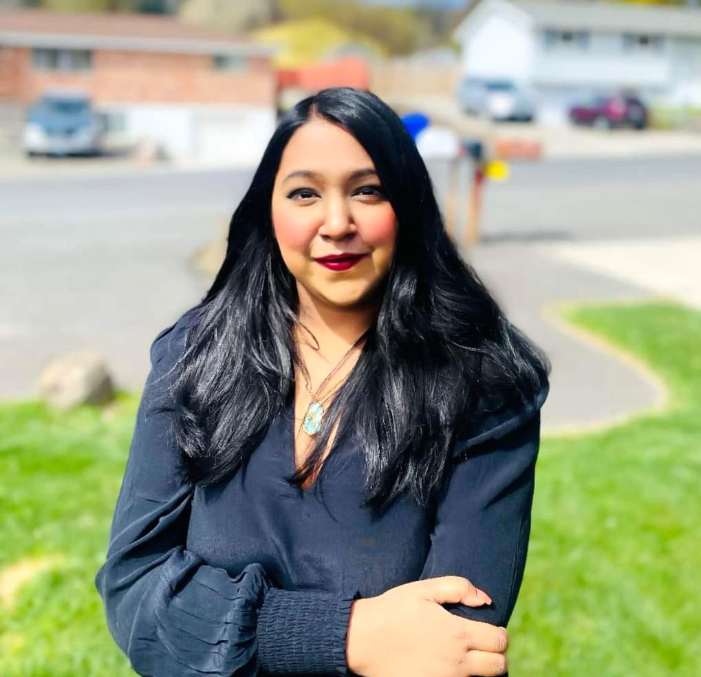 A woman with long black hair and dark red lipstick standing outdoors on a sunny day, wearing a black jacket and a necklace, with a blurred background of a street and houses.