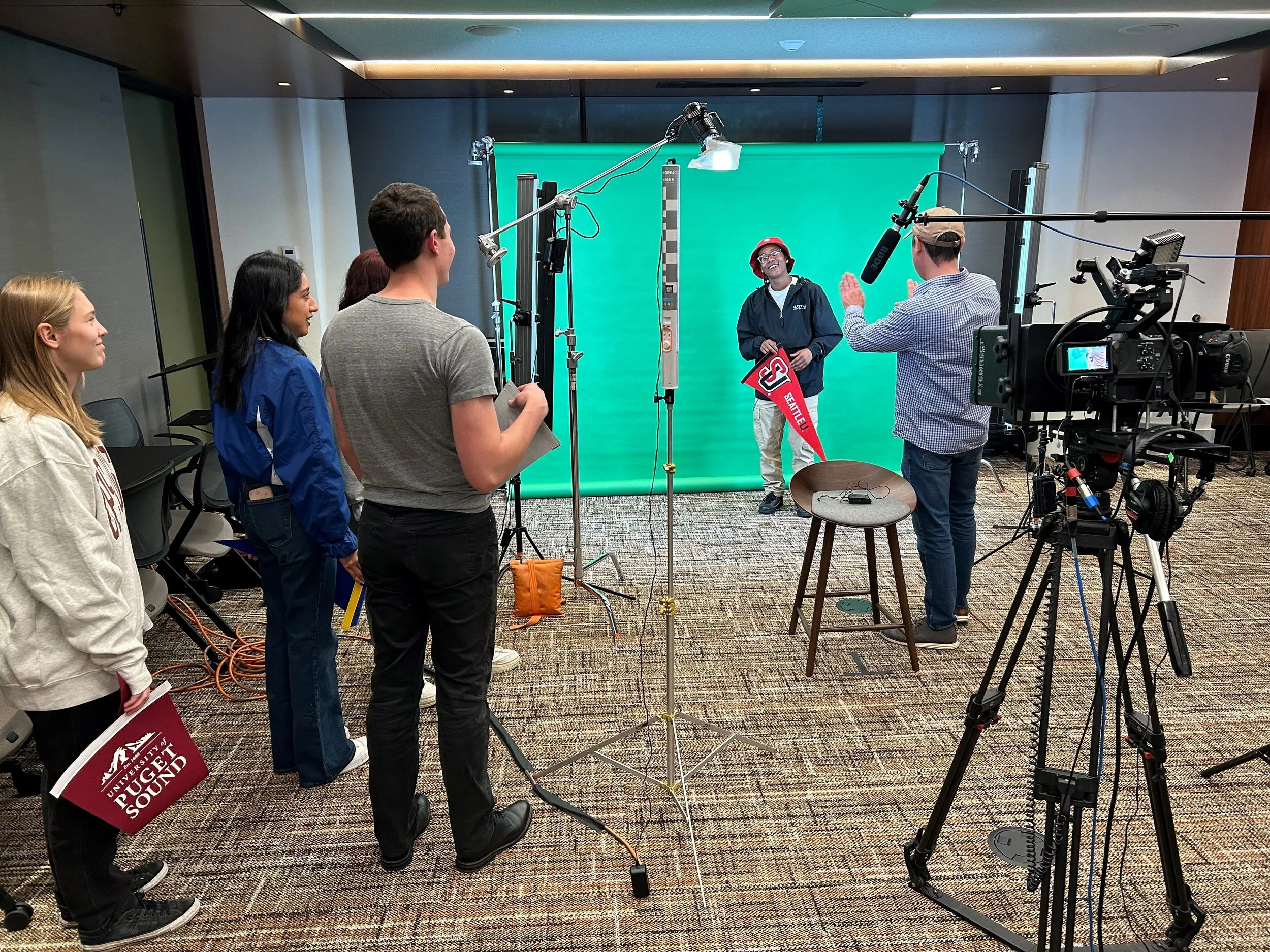 Group of people filming a video in a studio with green screen. One person is speaking, others are observing or holding notes. Equipment includes cameras, microphones, and lighting.