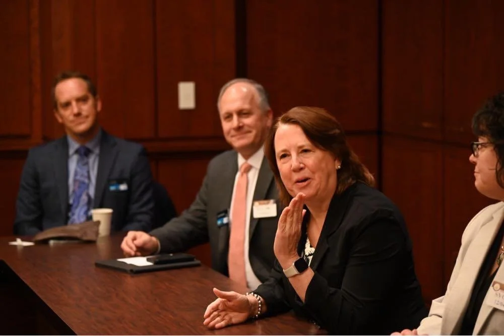 Four people sitting at a conference table engaged in discussion, with a woman speaking and others listening.