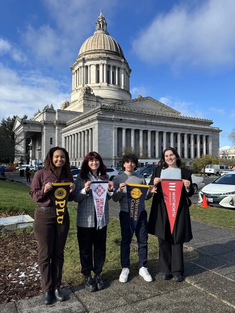 Four young women standing in front of the historic state capitol building, holding banners representing different universities.