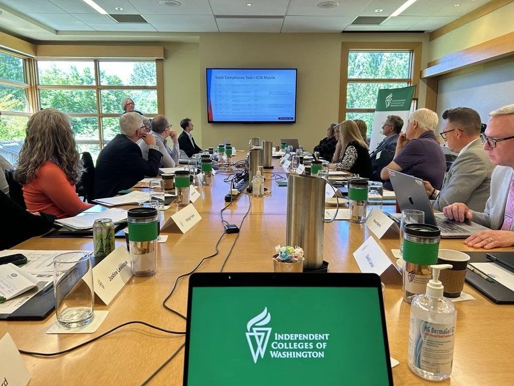A large conference room filled with people seated around a long table, watching a presentation on a wall-mounted screen. The table has laptops, water bottles, coffee cups, and name tags. A woman is standing at the front near the screen, speaking. Sunlight streams through large windows revealing greenery outside. A laptop in the foreground displays the logo of Independent Colleges of Washington.