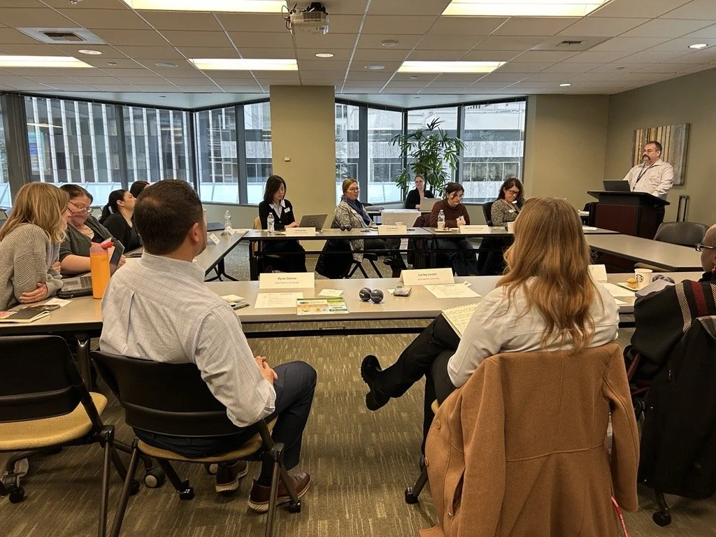 Business meeting in a conference room with multiple people seated at tables and a man standing at a lectern.
