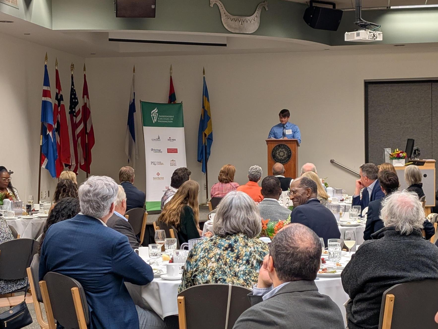 A young man giving a speech at a podium during a formal event with round tables filled with attendees in business attire, and flags of various countries displayed behind him, at Pacific Lutheran University.