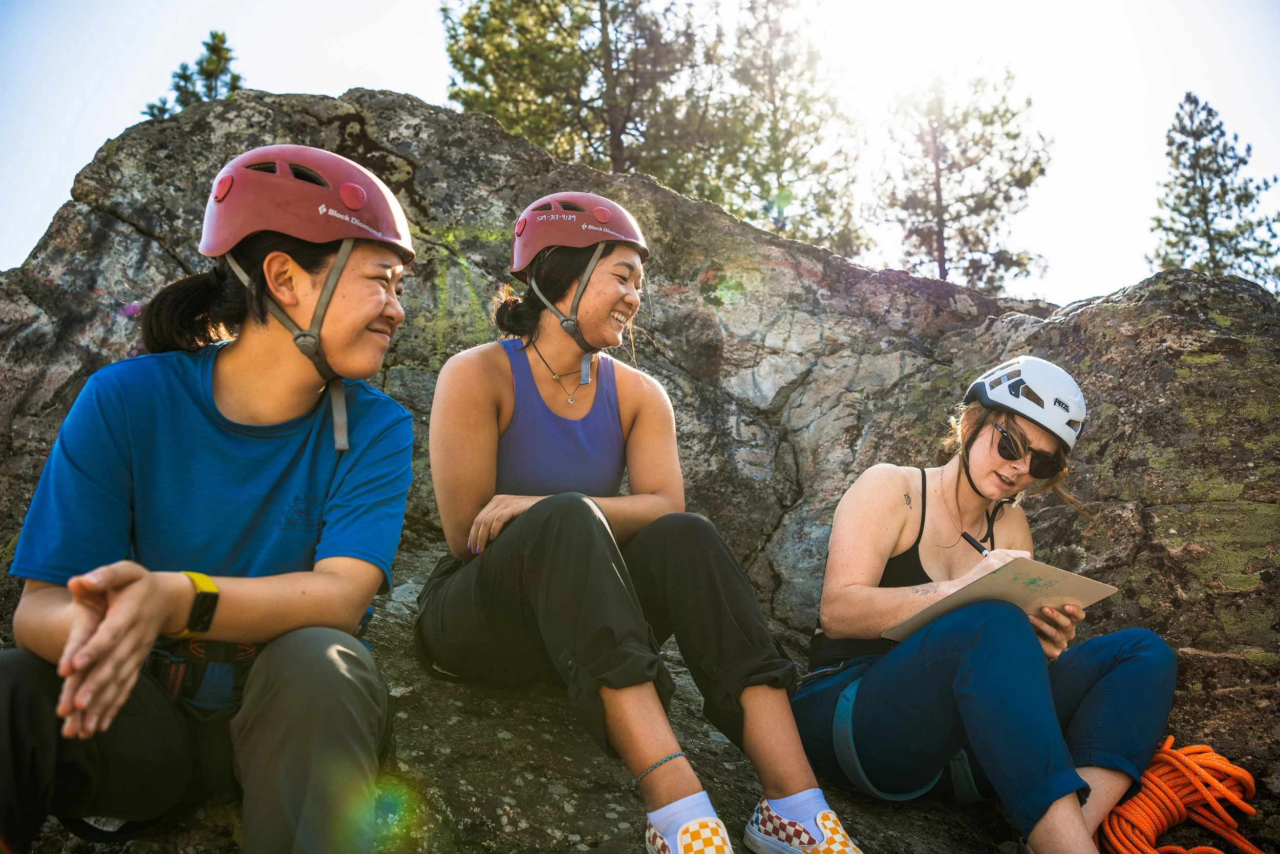 Three women with helmets sitting on rocks outdoors, smiling and writing on a notepad.