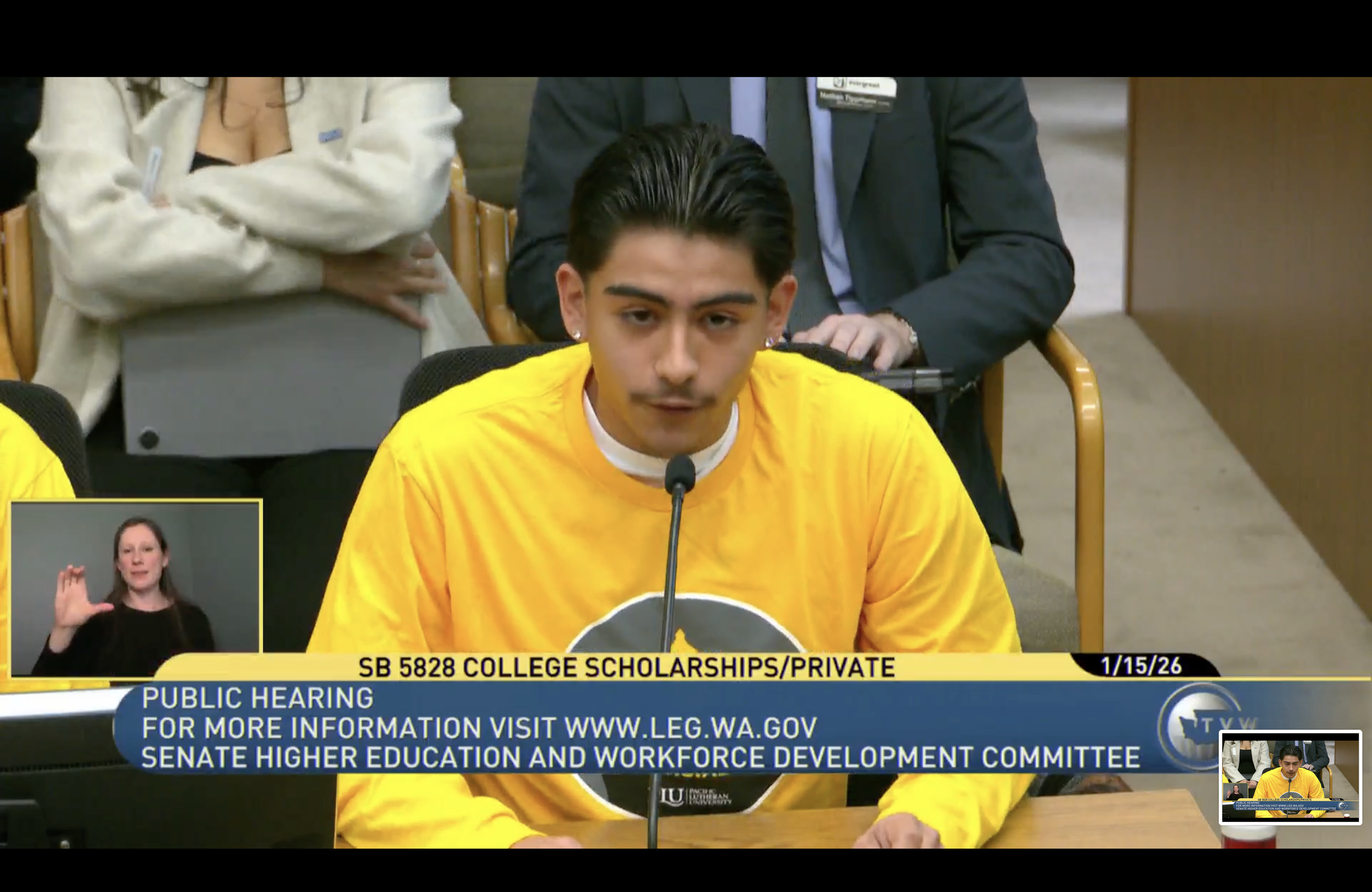 A young man wearing a yellow sweatshirt speaks into a microphone at a public hearing, with a woman and a man in suits behind him, and a sign about college scholarships.