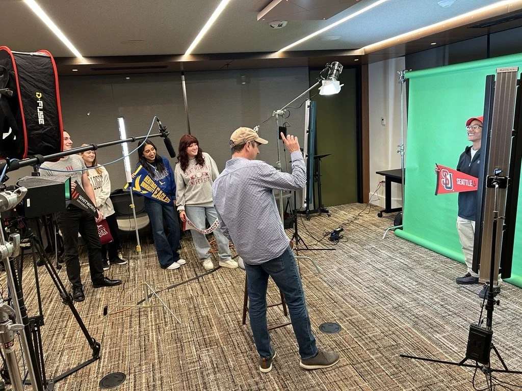 Group of five women standing in line for photo shoot with a man in front of a green screen, with professional lighting and camera equipment. The woman at the end holds a red