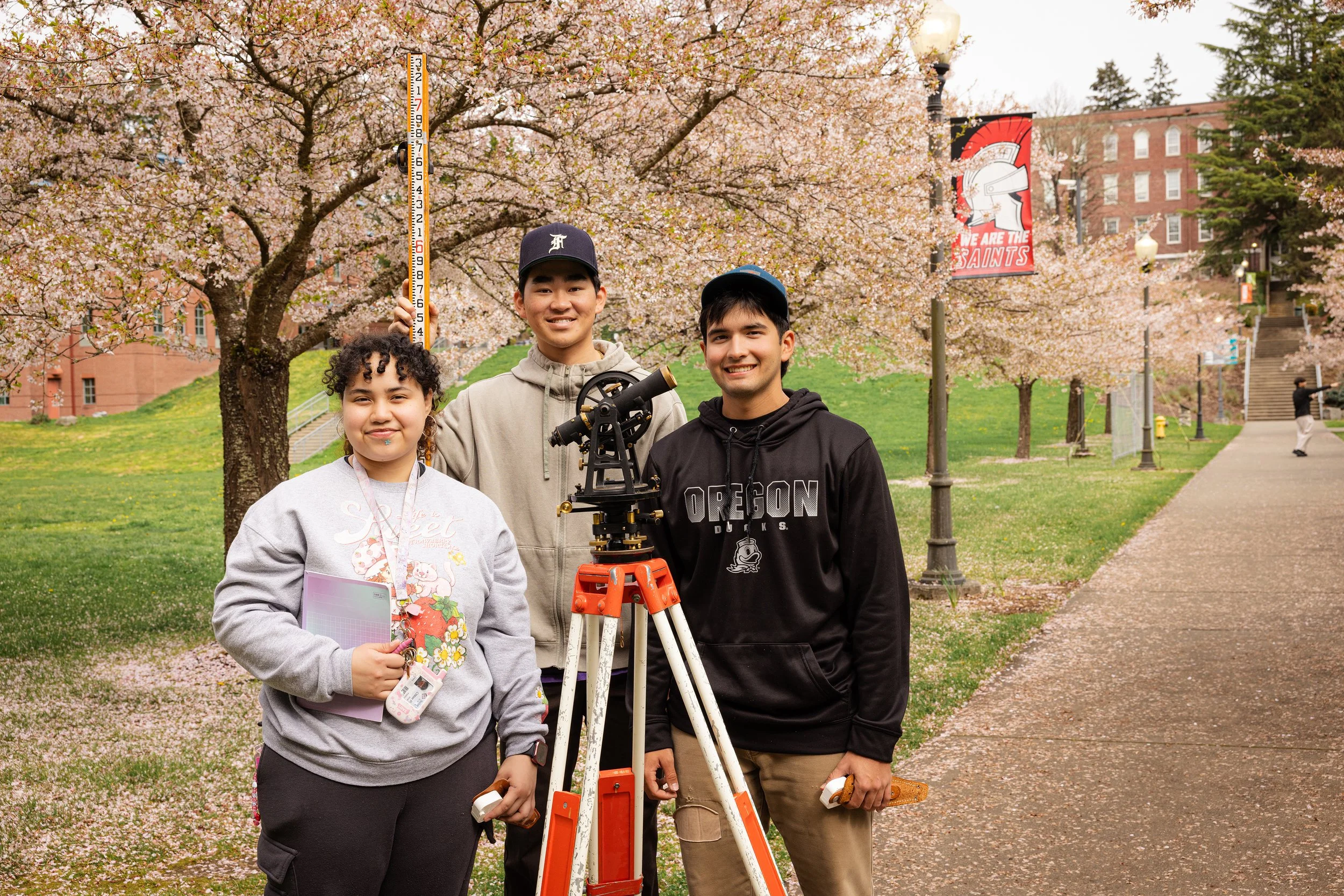 Three young people standing outdoors during cherry blossom season, with a cherry tree in full bloom behind them. One person holds a survey rod and a theodolite, possibly for measurement or surveying work. They are smiling and dressed casually, with a graduation cap on one person. There is a banner in the background reading 'We Are The Saints', and other people walking nearby on a path.