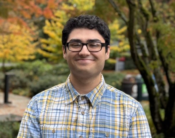 Young man with black curly hair and glasses, wearing a yellow and blue plaid shirt, standing outdoors with autumn foliage in the background.