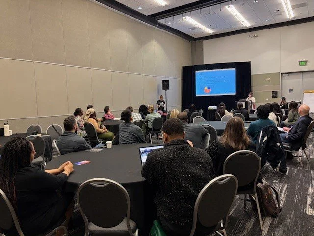 People sitting at round tables attending a presentation in a conference room with a speaker and a projection screen.