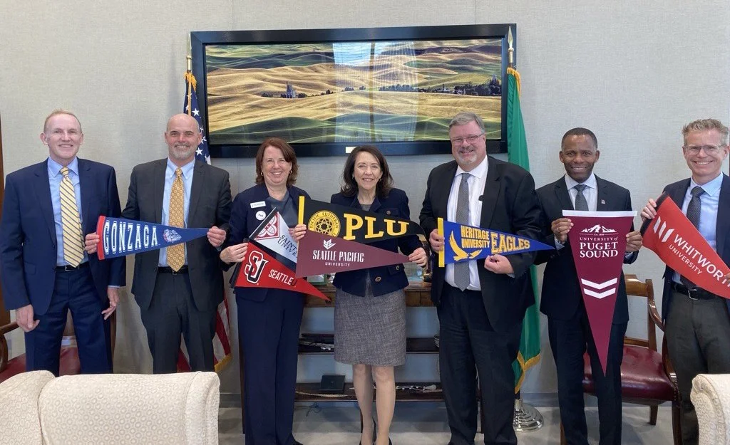 Group of seven professionals holding college and university pennants, standing indoors in front of a screen displaying a landscape with rolling hills, with American and state flags behind them.