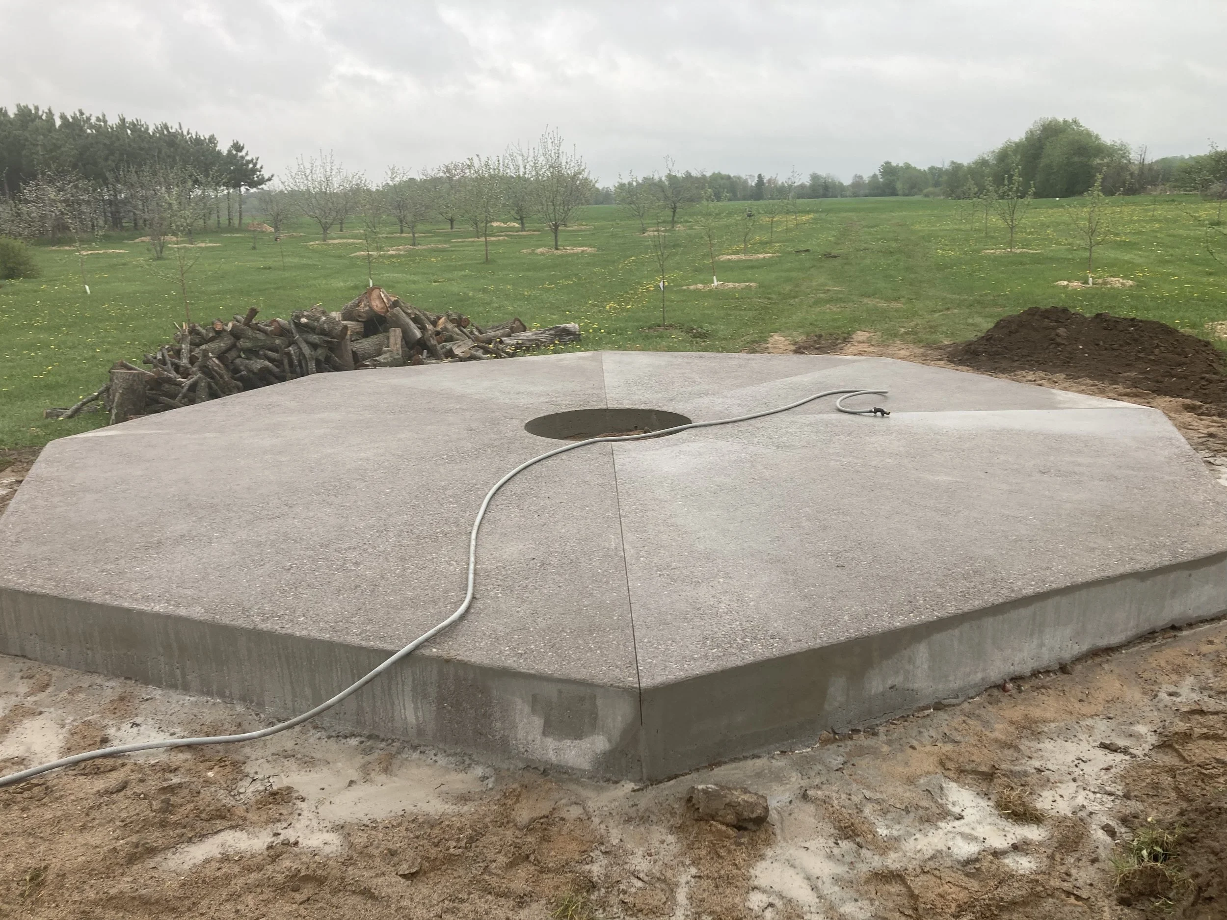 Concrete slab with a circular hole in the center, connected to an extension cord, outdoors in a grassy field with trees and a pile of firewood in the background.