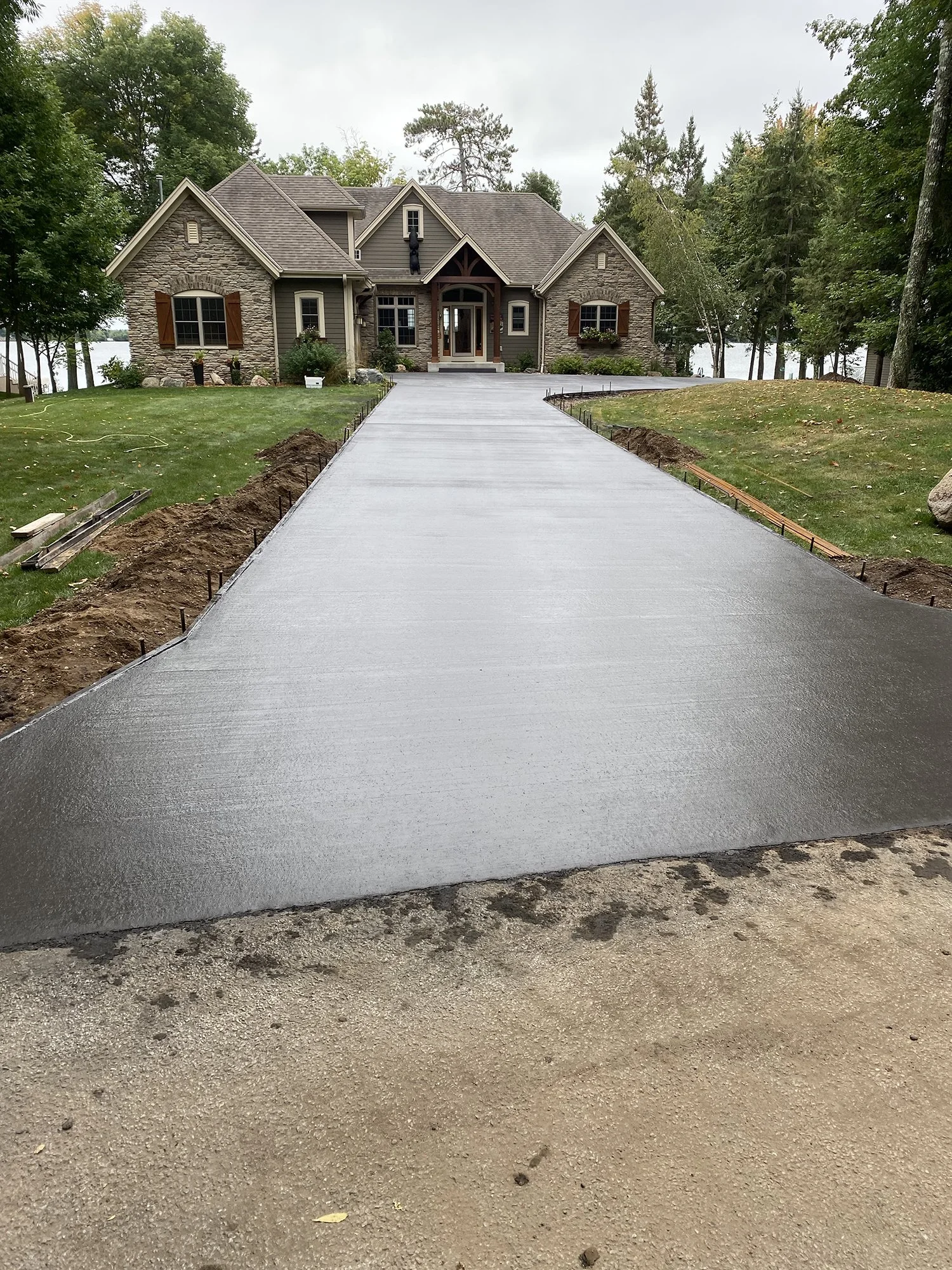 Freshly poured concrete driveway leading to a large house with a stone and siding exterior, surrounded by trees and grass.