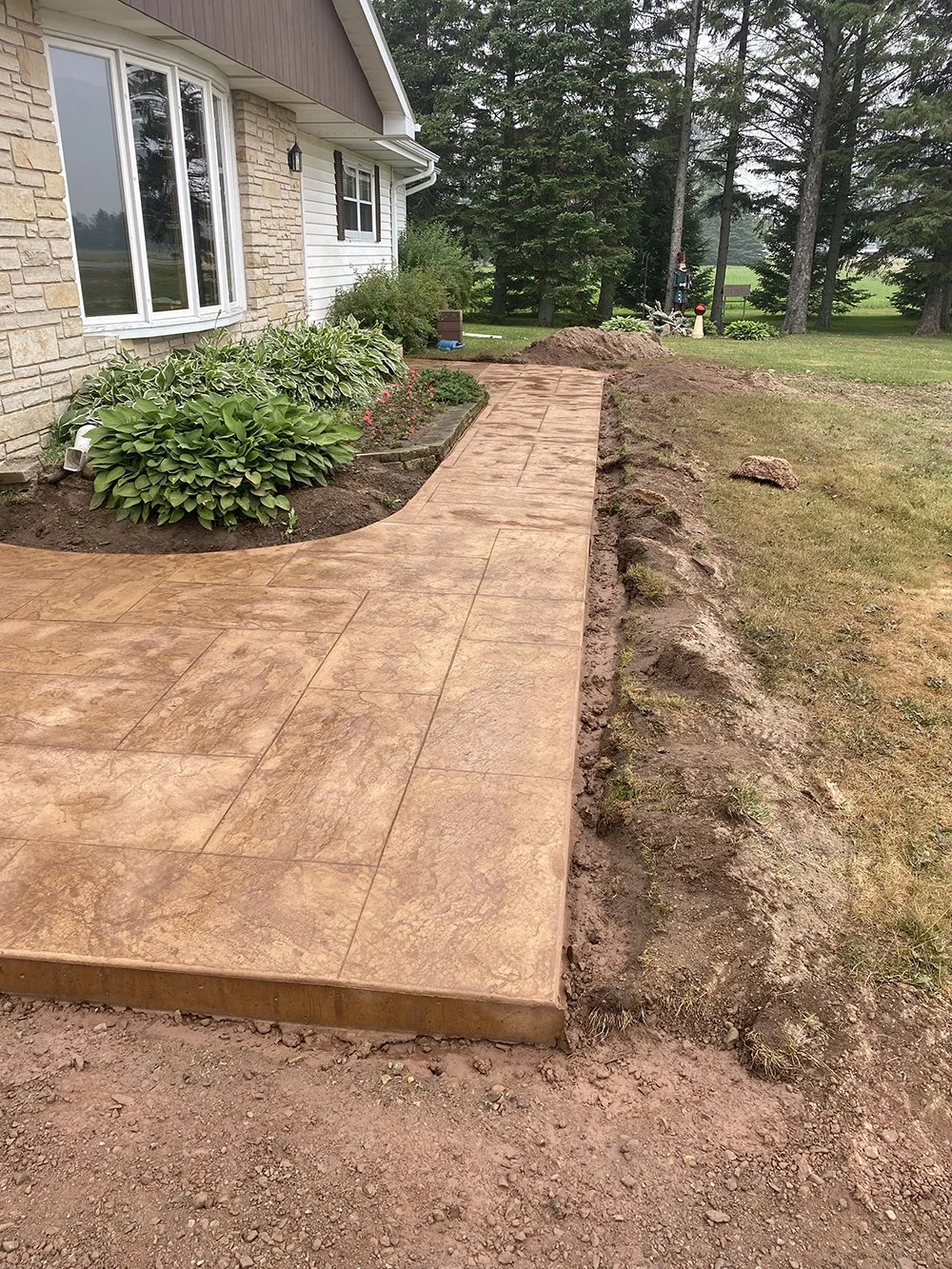 Photo of a freshly laid concrete sidewalk edges, with an ongoing garden landscaping project in front of a house with large windows, surrounded by green plants and trees.