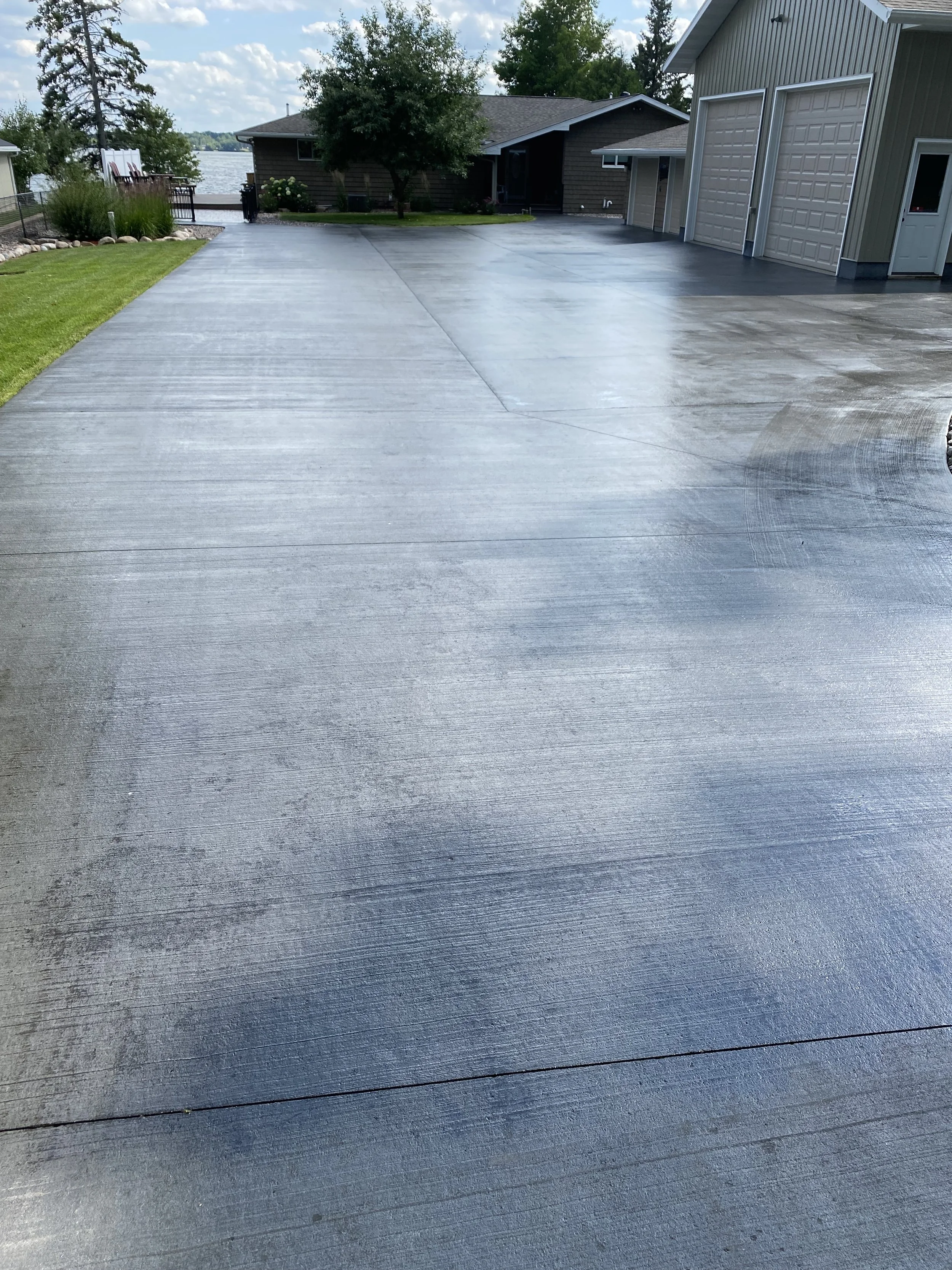 Clean, wet concrete driveway in front of a house with a lawn and trees, overlooking a body of water with a cloudy sky.