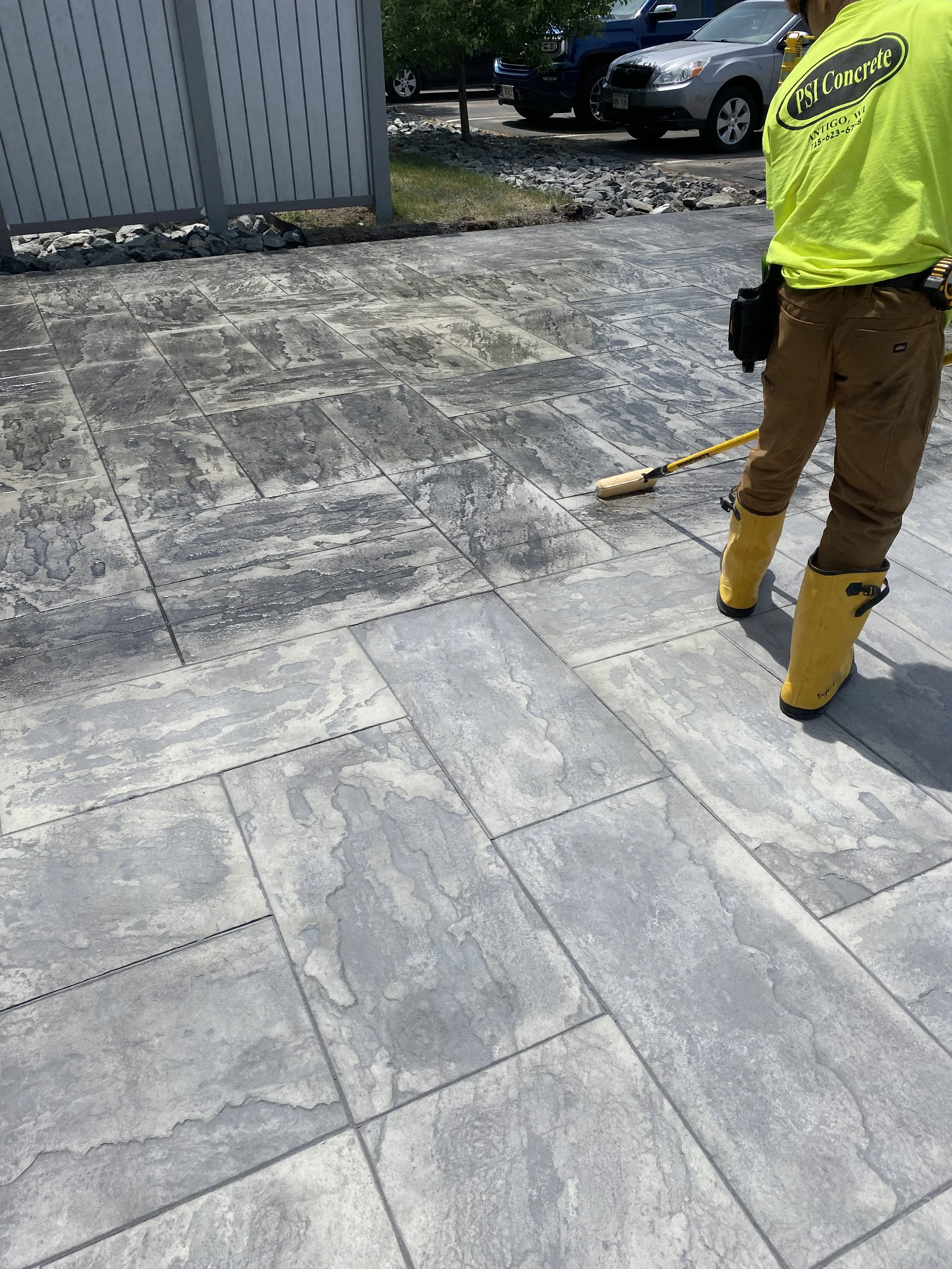 Worker cleaning or sealing a concrete outdoor patio with a brush, wearing yellow rubber boots and a neon yellow T-shirt with 'PSI Concrete' printed on the back