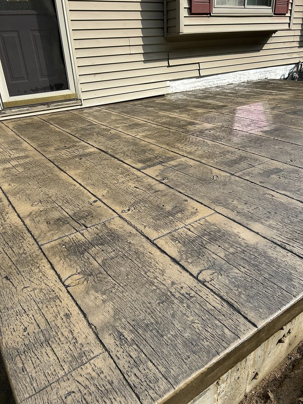 Close-up view of a weathered wooden porch or deck outside a house with beige vinyl siding, a partially visible door, and a window with red shutters.