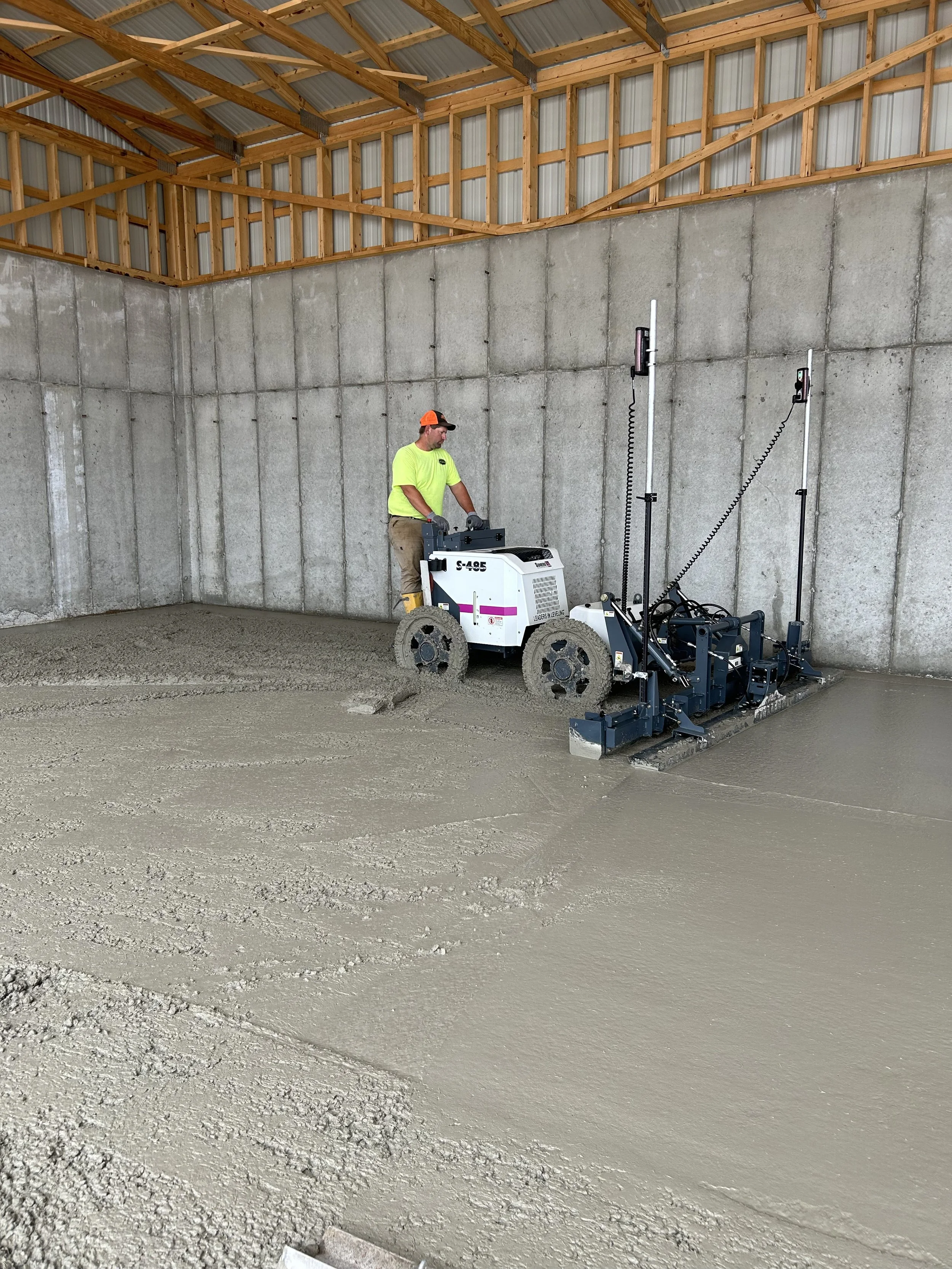 A worker in a neon yellow shirt and tan pants operates a concrete finishing machine on wet concrete inside a building under construction with exposed concrete walls and a wooden roof structure.