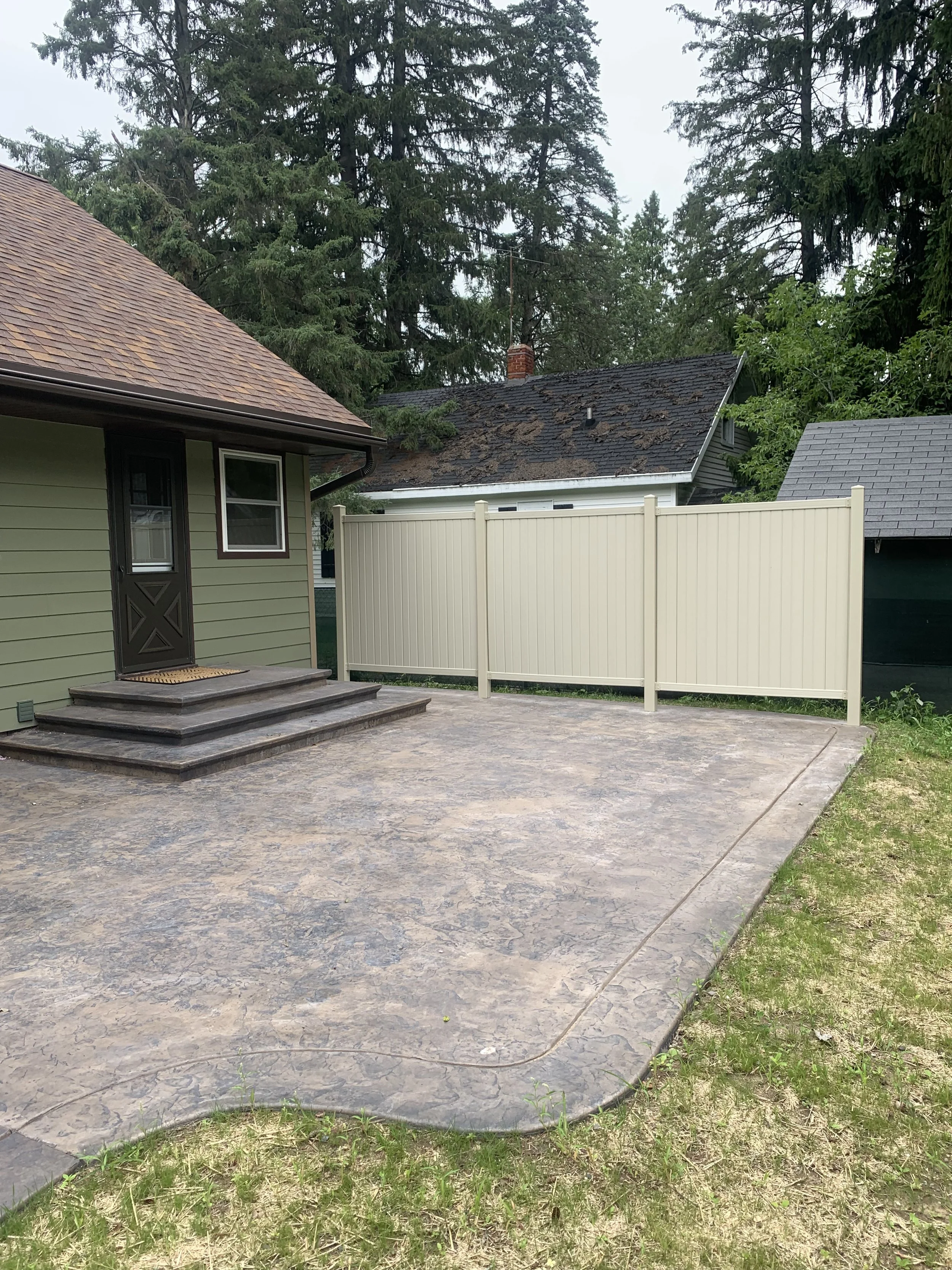 A backyard patio with a stamped concrete surface leading up to the steps of a house with a brown shingled roof and green siding, and a white privacy fence in the background, with trees in the yard.