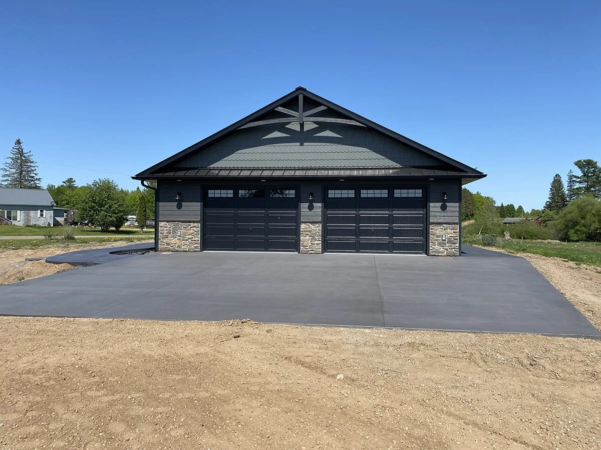 A newly paved black asphalt driveway leading to a black garage with two doors, surrounded by a rural landscape with green trees and a clear blue sky.