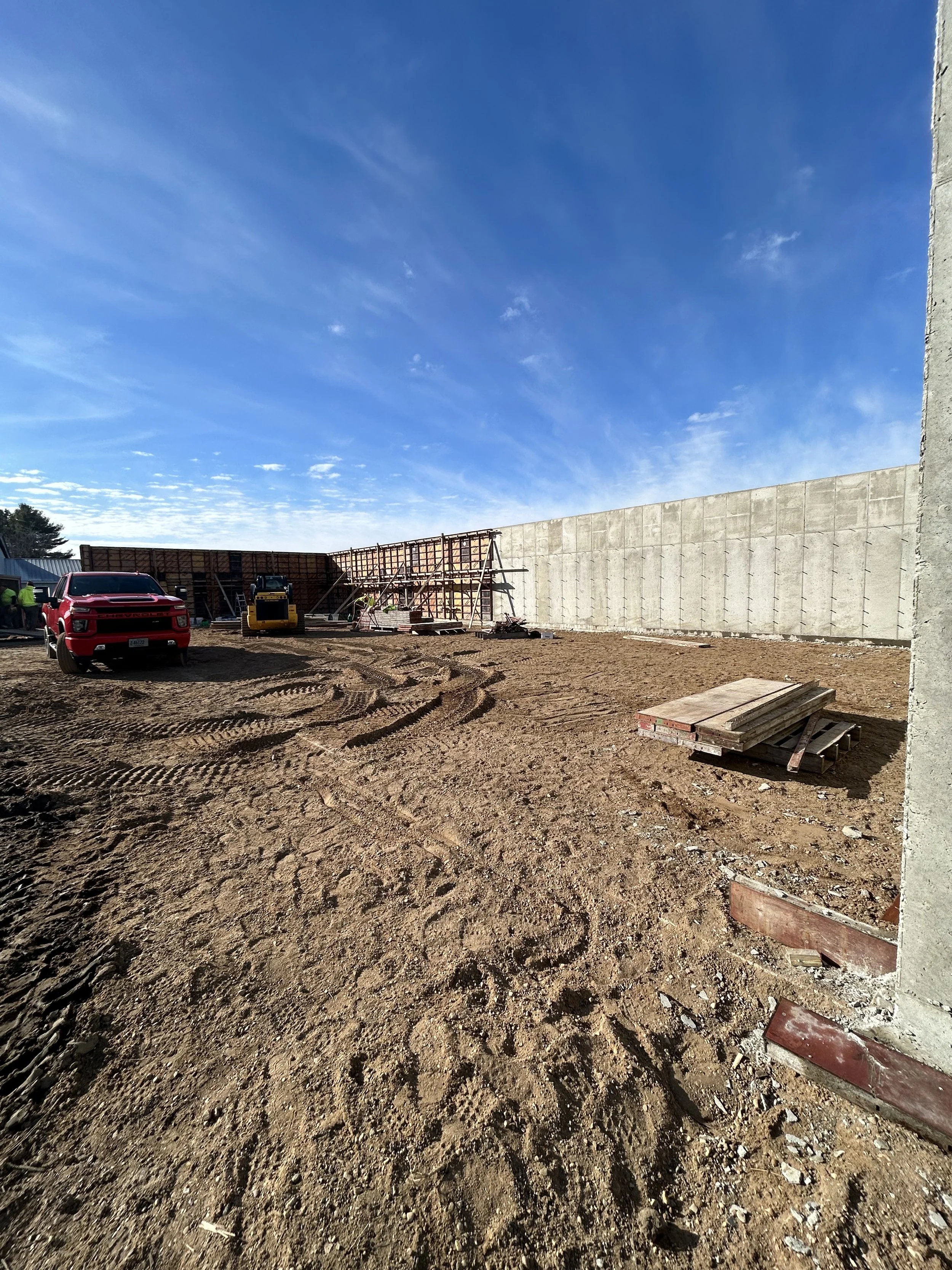 Construction site with dirt ground, construction equipment, vehicles, and wooden planks under a blue sky with clouds.